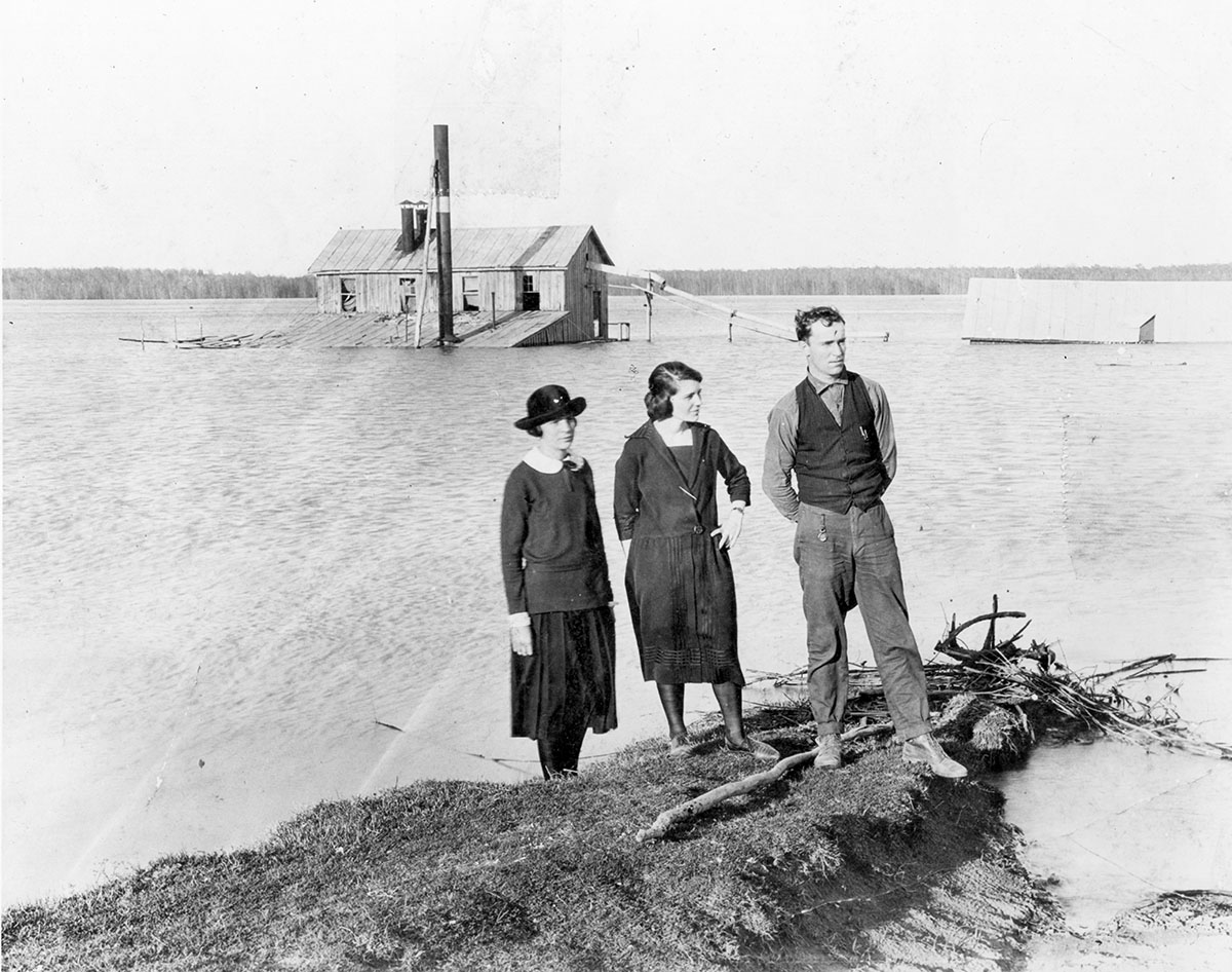 Two women and a man stand on a levee surrounded by water; house in background